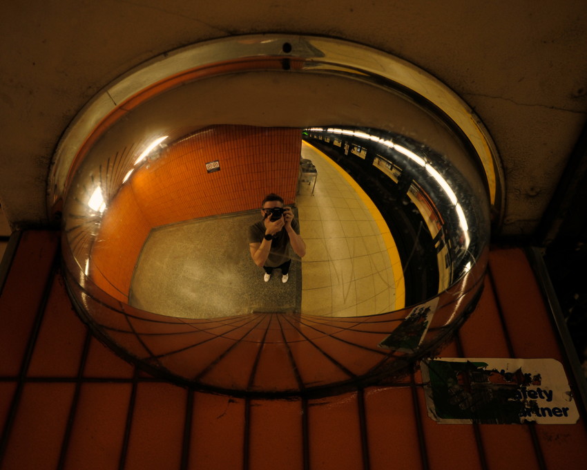 Convex mirror reflecting photographer on subway platform, orange tiled wall, yellow platform edge, curved fisheye view.