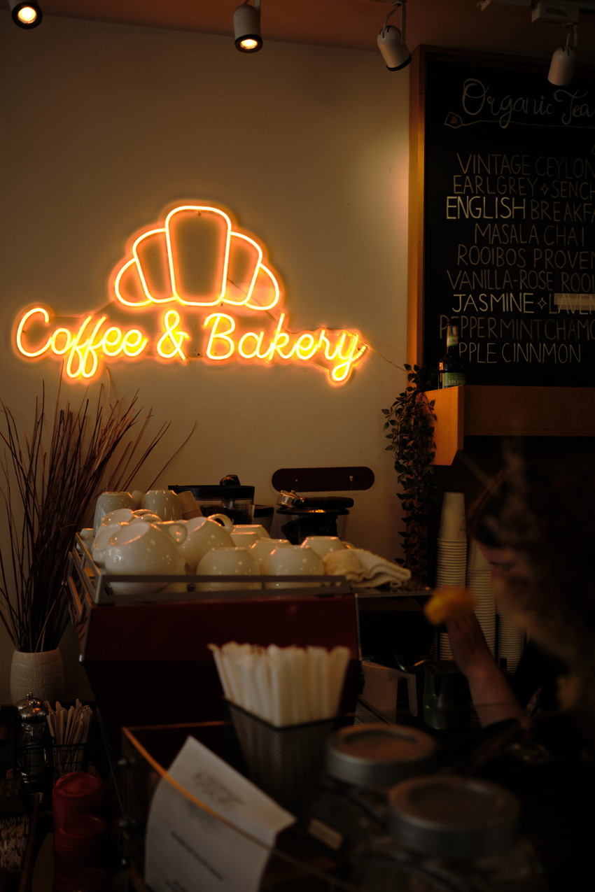 Interior café counter with neon croissant logo and 'Coffee & Bakery' sign, stacked white cups, espresso machine, chalkboard menu.