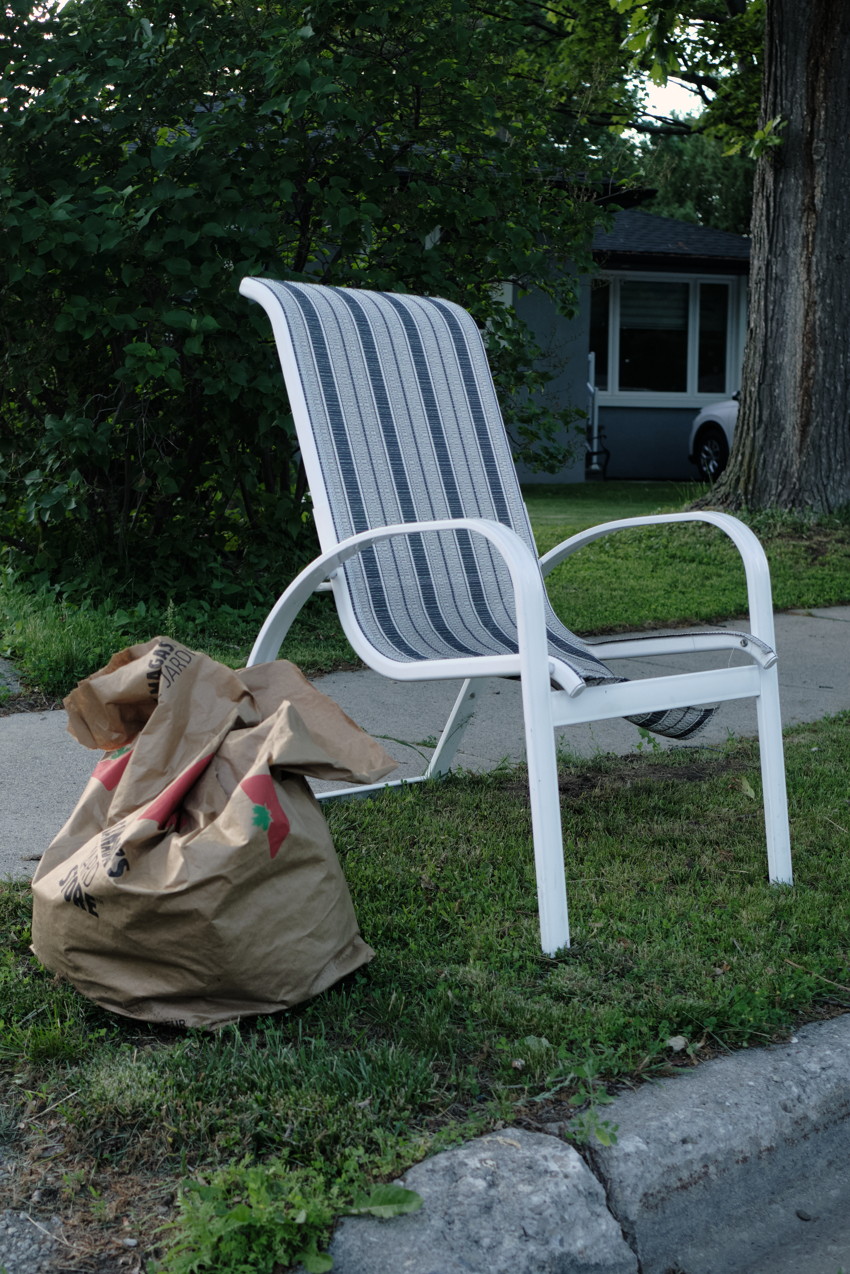 White metal outdoor chair with blue-striped seat on lawn near curb; brown yard-waste paper bags beside it.