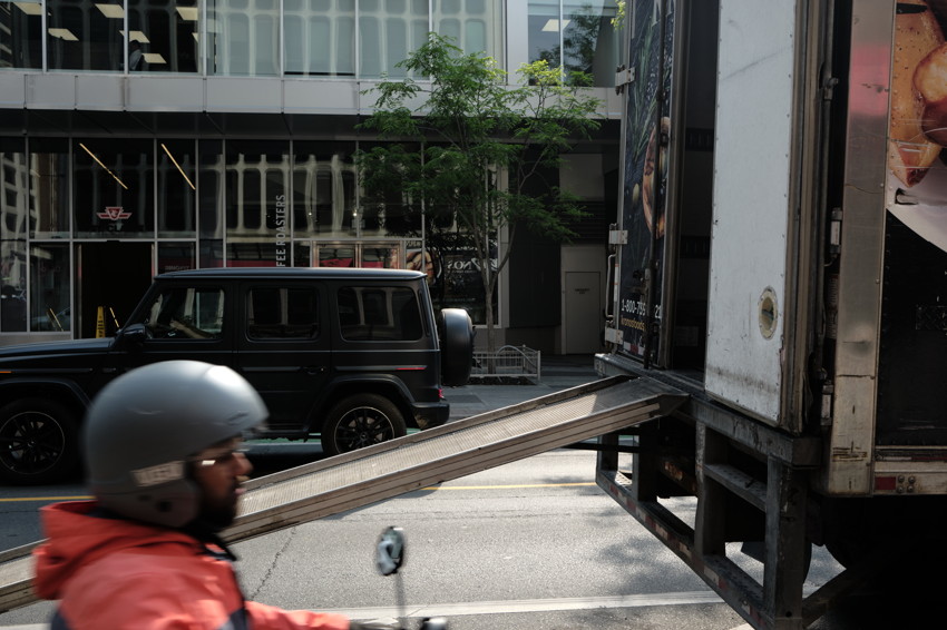 Blurred helmeted motorcyclist foreground; ramp into open delivery truck right; black SUV and glass storefront behind.