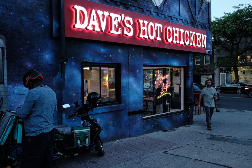Dave's Hot Chicken storefront at night: red neon sign, galaxy-painted facade, delivery scooter, passerby.