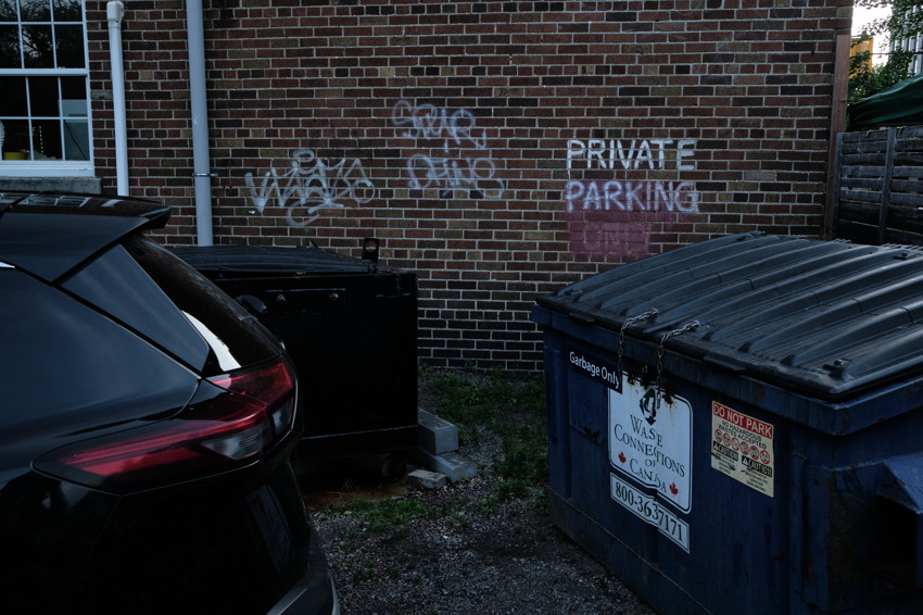 Rear of parked car, two dumpsters chained beside graffiti-covered brick wall reading 'Private Parking', gravel ground.