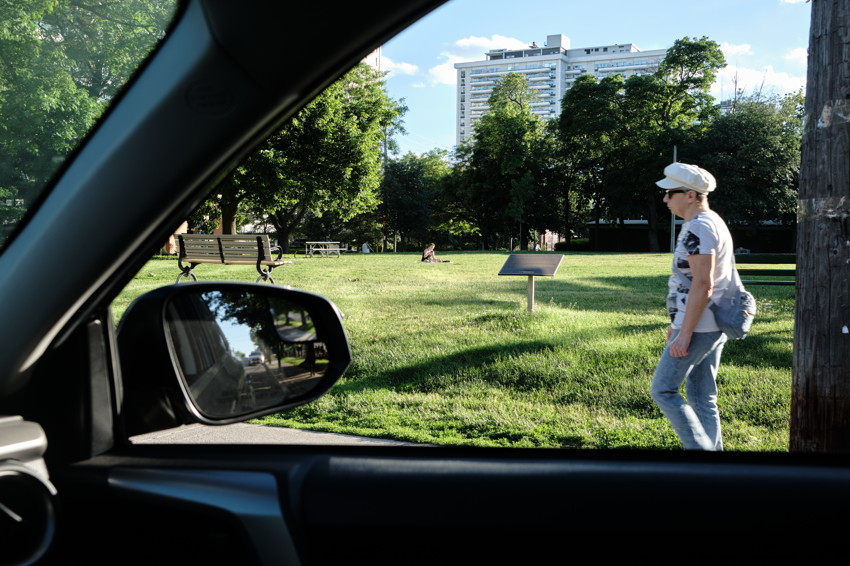 Park lawn framed by car window; side mirror reflection of street; person walking right, high-rise behind.