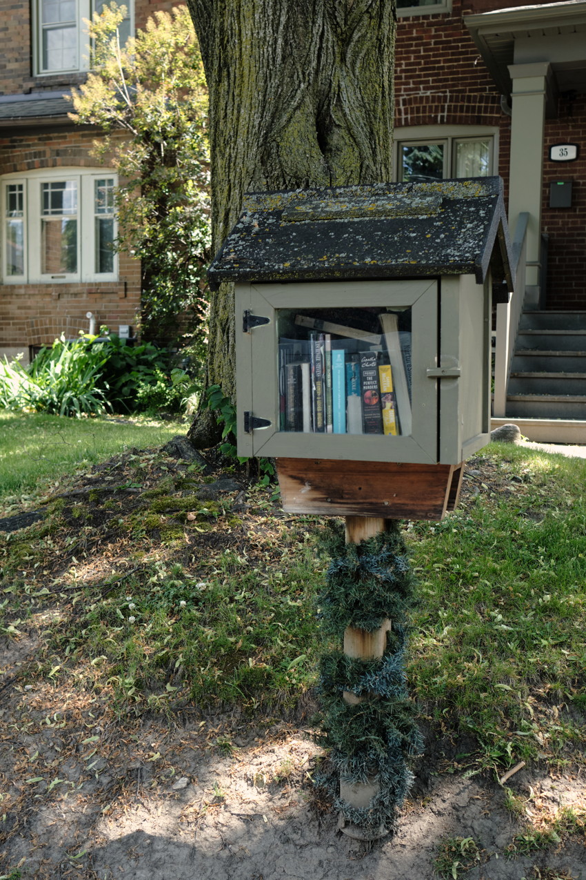 Small wooden free library box on a post with books, in front of a tree and brick house.