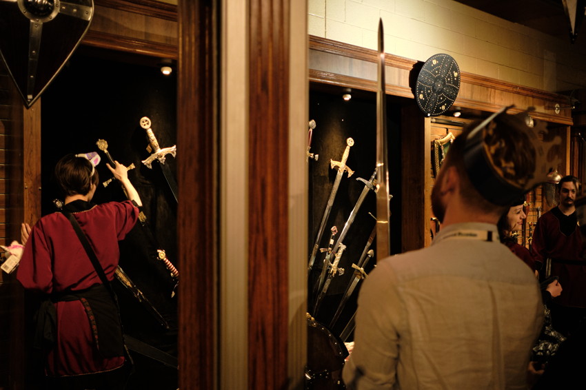 Glass museum case with multiple swords arranged fanwise; visitors observe either side of wooden divider.