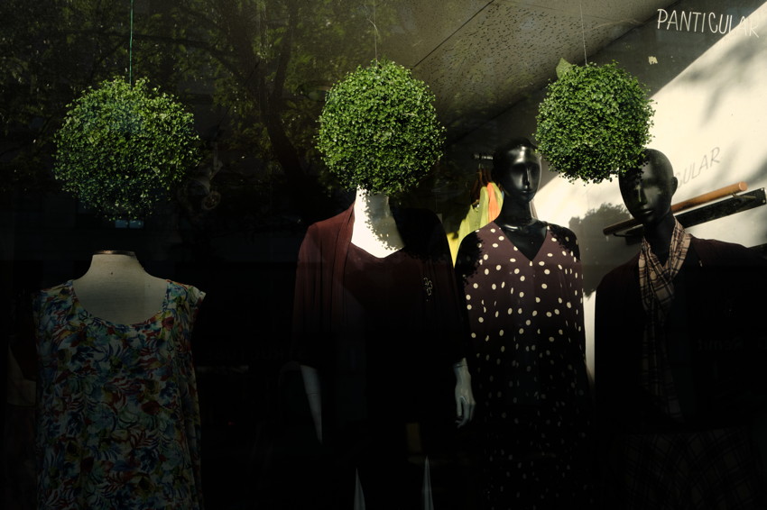 Shop window with four mannequins; three hanging spherical green plants positioned above their heads, clothing on display.