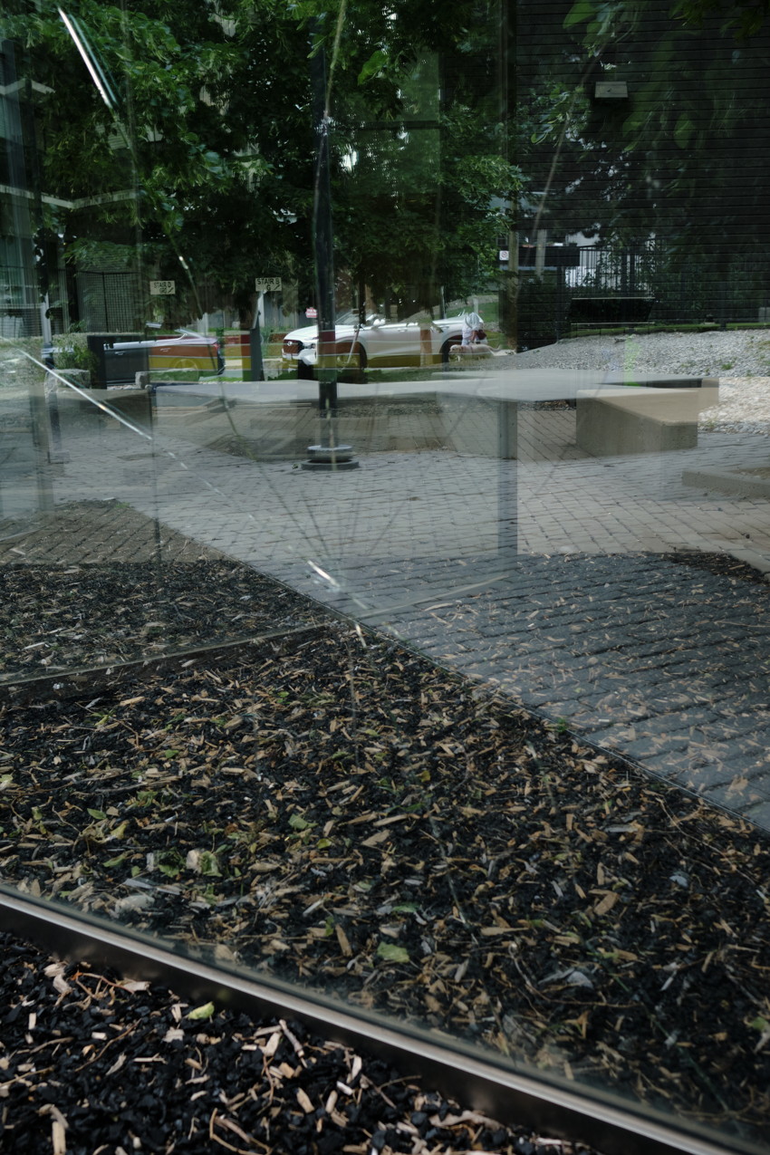 Shattered glass pane reflecting trees and parked car; foreground wood chips, paved plaza and low concrete benches.