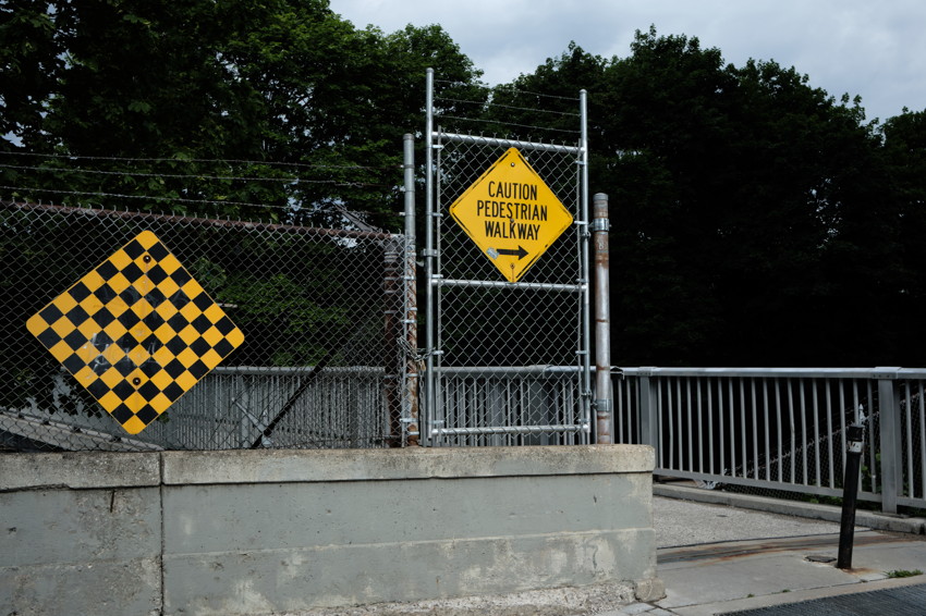 Chain-link fence, concrete barrier, yellow checkered sign and Caution Pedestrian Walkway sign by bridge railing.