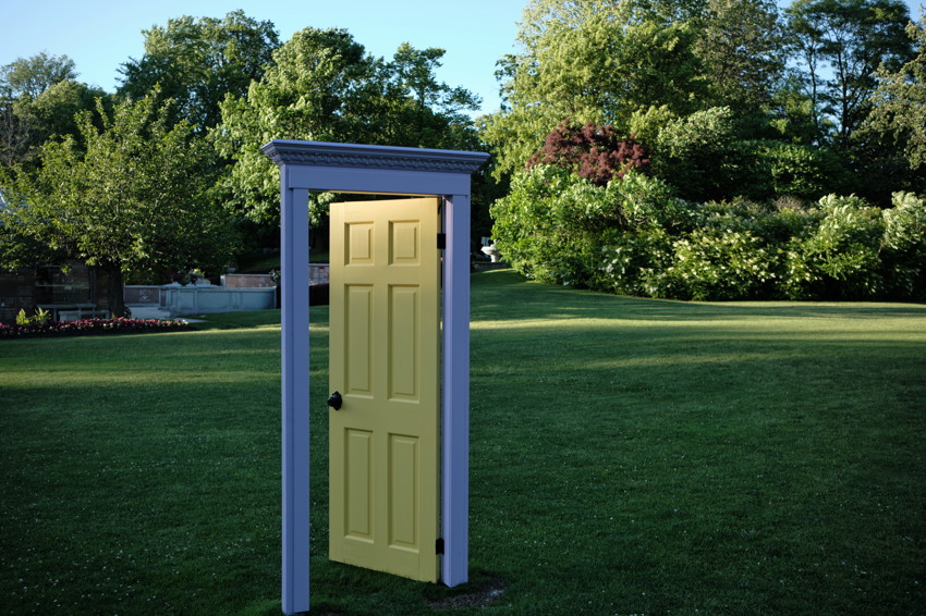 Freestanding door frame with partially open yellow door standing on lawn, trees and shrubs in background.