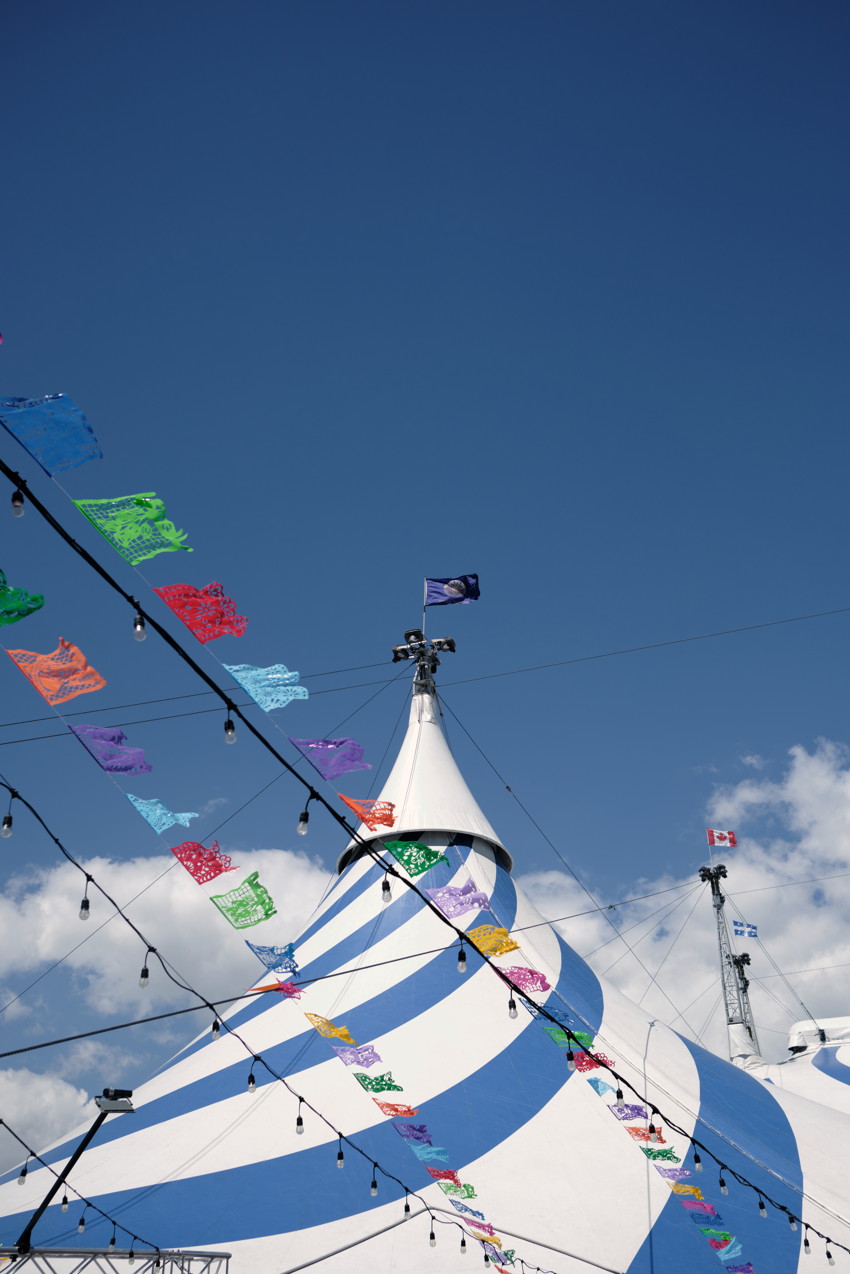Blue-and-white striped circus tent peak with colorful papel-picado banners and string lights against clear blue sky.