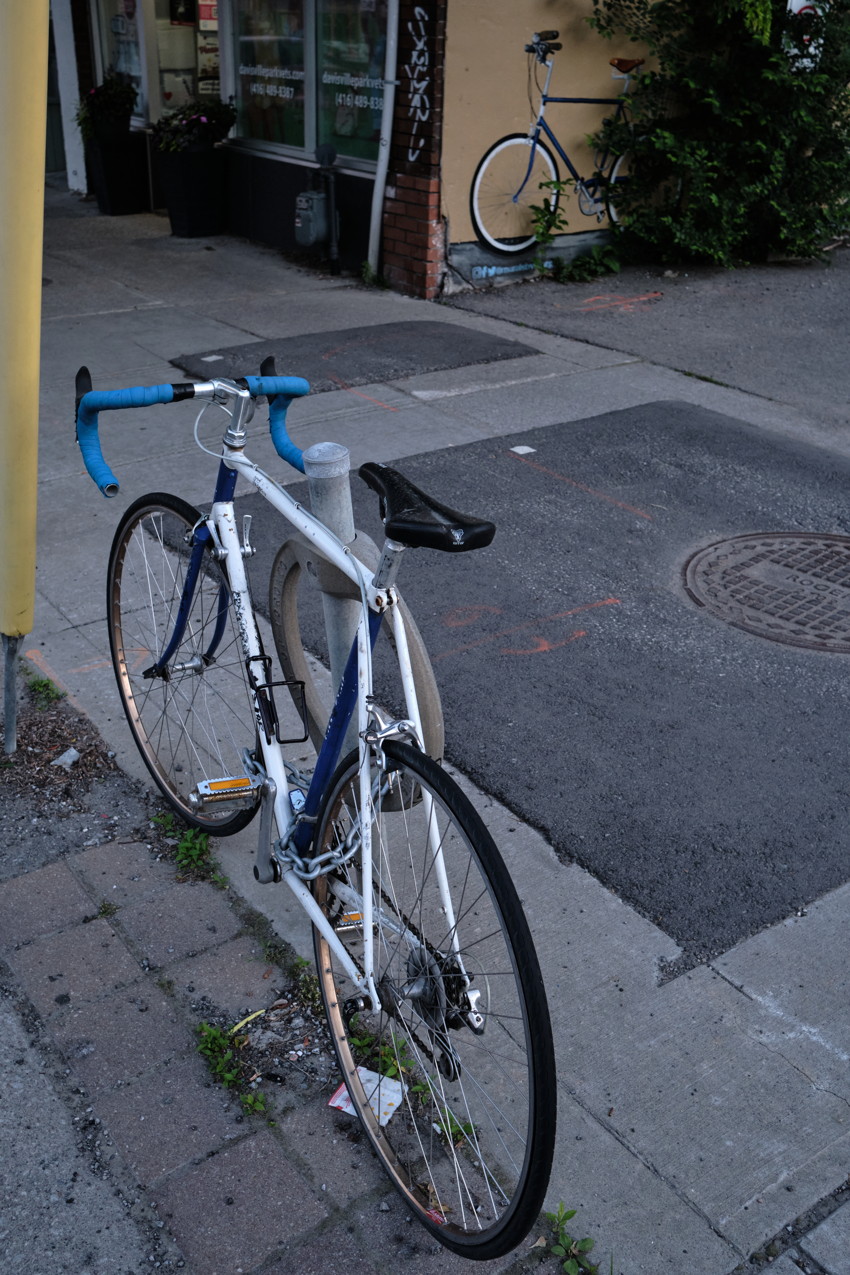 White-and-blue road bicycle chained to a short bollard on a cracked sidewalk, storefront and second painted bike on wall in background.