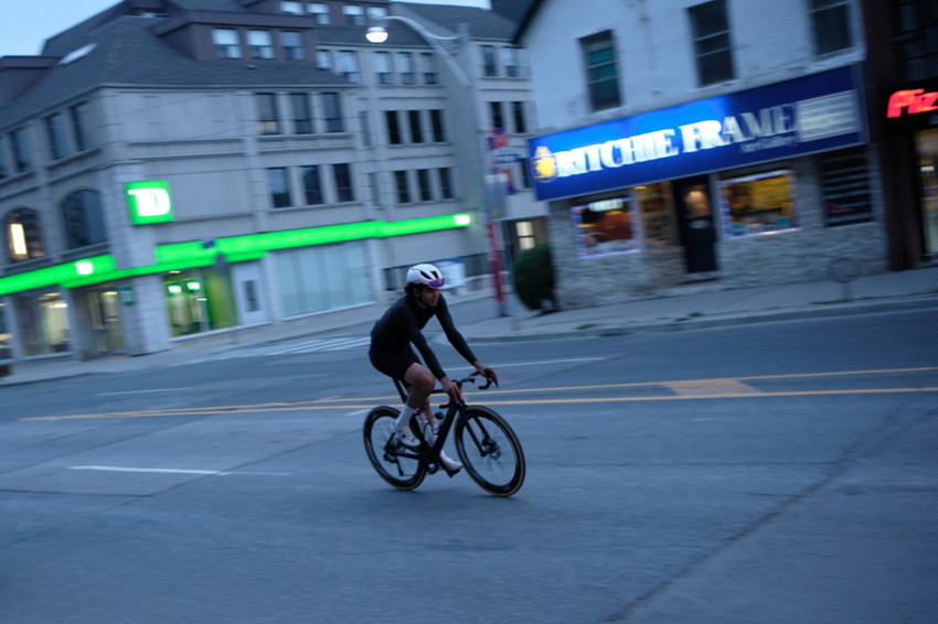 Cyclist in black kit and white helmet leaning into turn on city street at dusk, storefronts and bank in background.