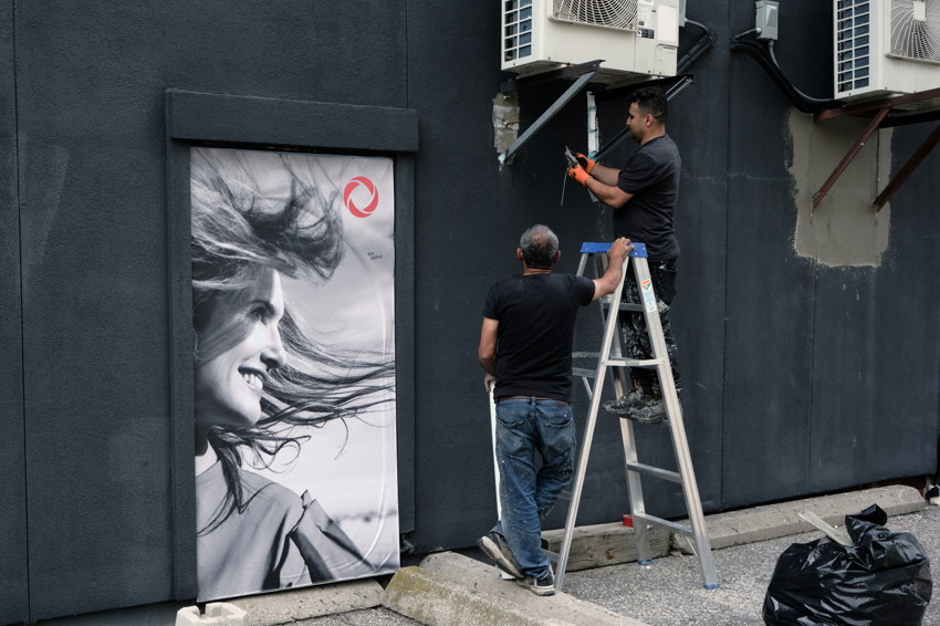 Two men, one on ladder, repairing wall-mounted air conditioners beside large black-and-white portrait poster.