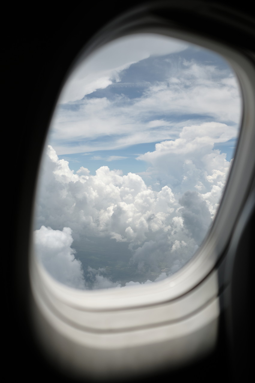 Airplane window framing layered cumulus clouds and blue sky above them.