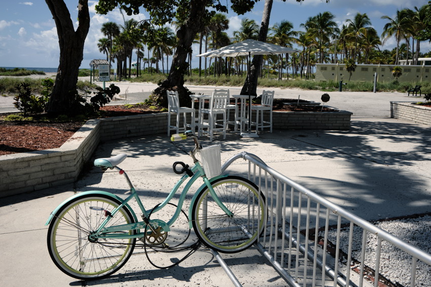 Mint-green bicycle locked to rack; white table and umbrella with palm trees and beach visible beyond.