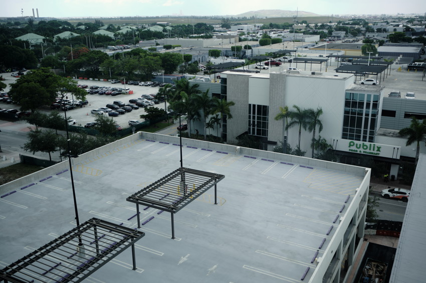 Aerial view of empty rooftop parking deck with metal shade frames, lampposts; Publix and city buildings in background.
