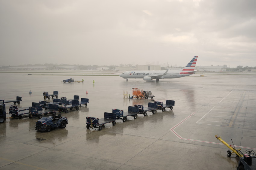 Wet airport apron with American Airlines jet taxiing, lined baggage carts and ground vehicles under overcast sky.