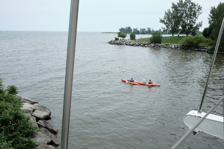 Two people in orange kayaks on a lake, rocky shore and grassy park with trees, metal railing foreground.