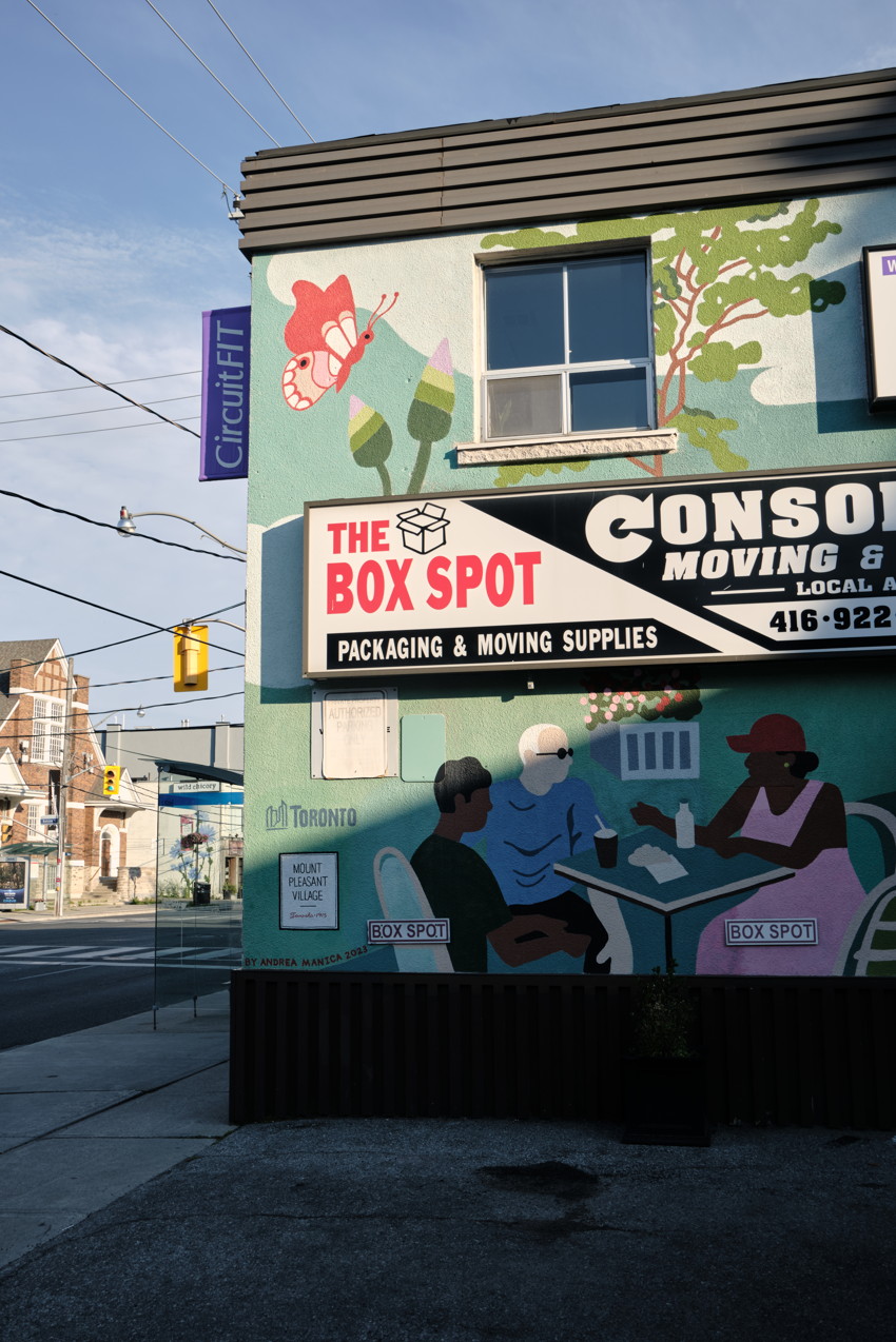 Storefront mural with 'The Box Spot' sign, painted people at table, tree and butterfly, street intersection.