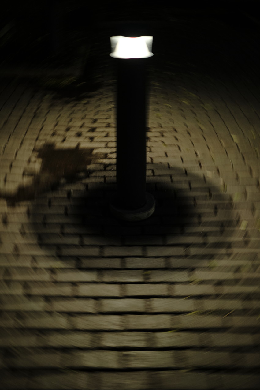 Short bollard light centered on brick pavement at night, circular illuminated area and radial motion blur.