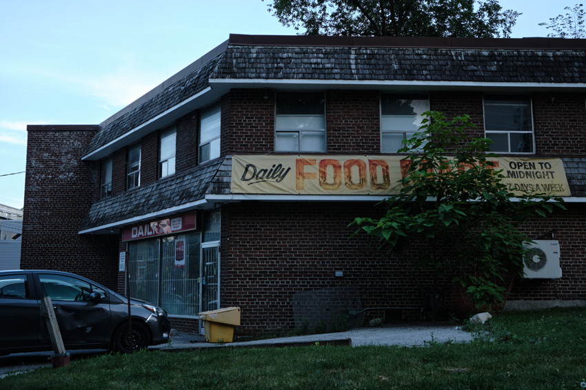Brick corner grocery with a faded Daily Food sign, barred entrance, parked car, and tree partly covering the facade.