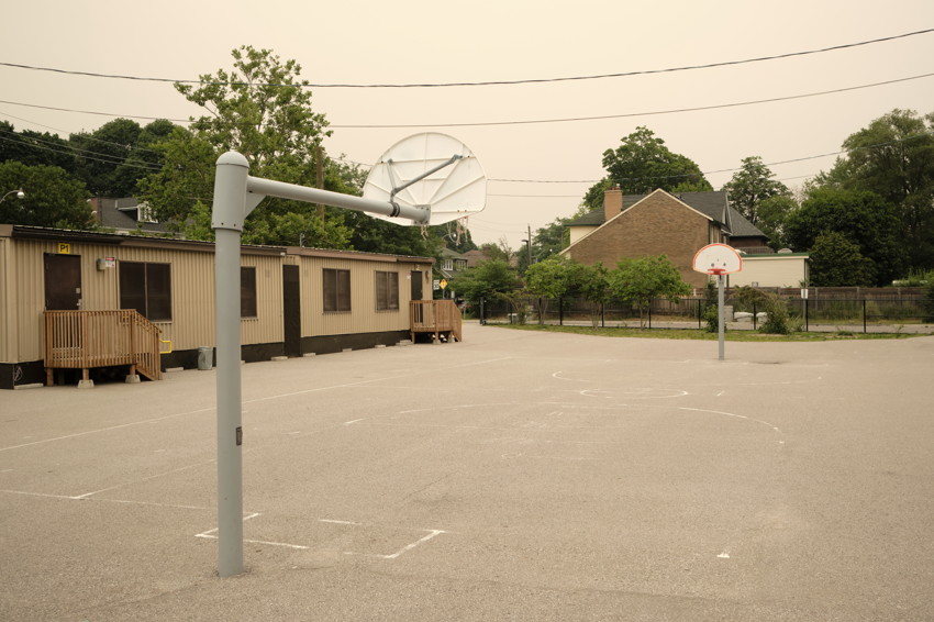 An empty asphalt schoolyard with two basketball hoops, a portable classroom, and nearby houses beneath a smoke-hazed sky.