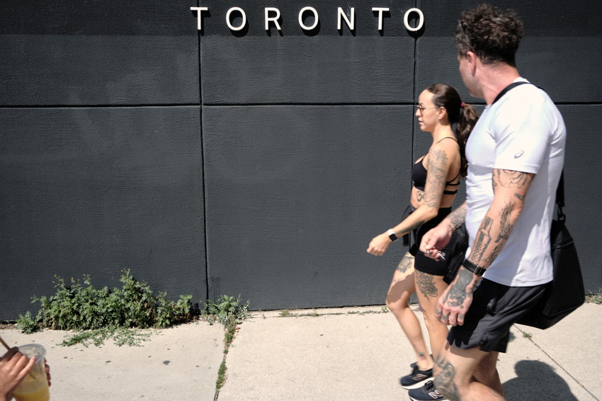Two pedestrians walk past a black wall with large white letters spelling TORONTO.
