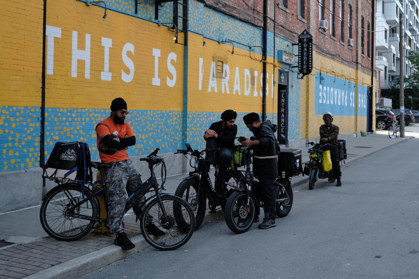 Four cyclists with cargo bicycles gather beside a mural-painted brick wall with 'THIS IS PARADISE' sign.