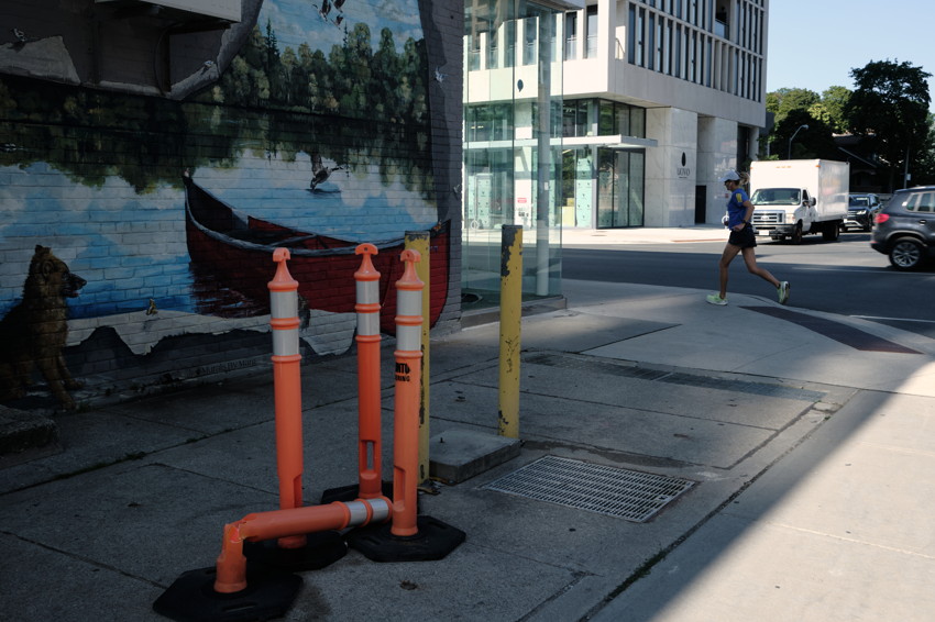 Orange traffic bollards on a sidewalk beside a mural of a boat, with a runner and vehicles on the street.
