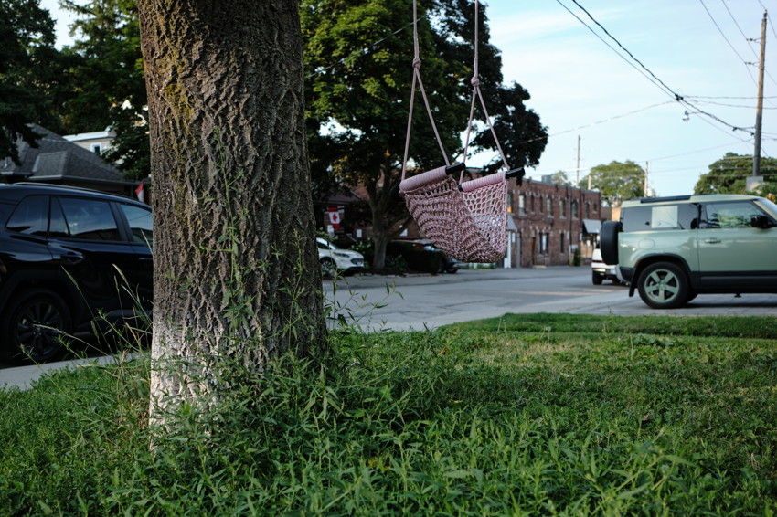 Pink woven swing hanging from a tree beside a residential street with parked cars and overhead utility lines.