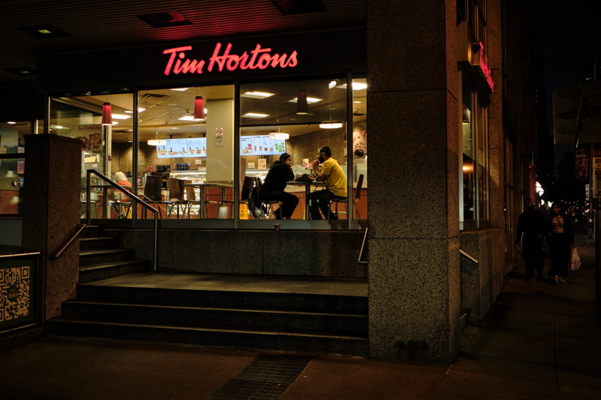 A Tim Hortons storefront at night, with two people seated inside near a stairway and a dark sidewalk.