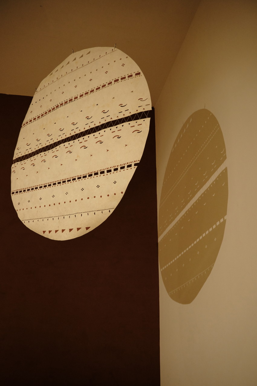 Oval patterned wall hanging with geometric bands casts a matching shadow on an adjacent beige wall.