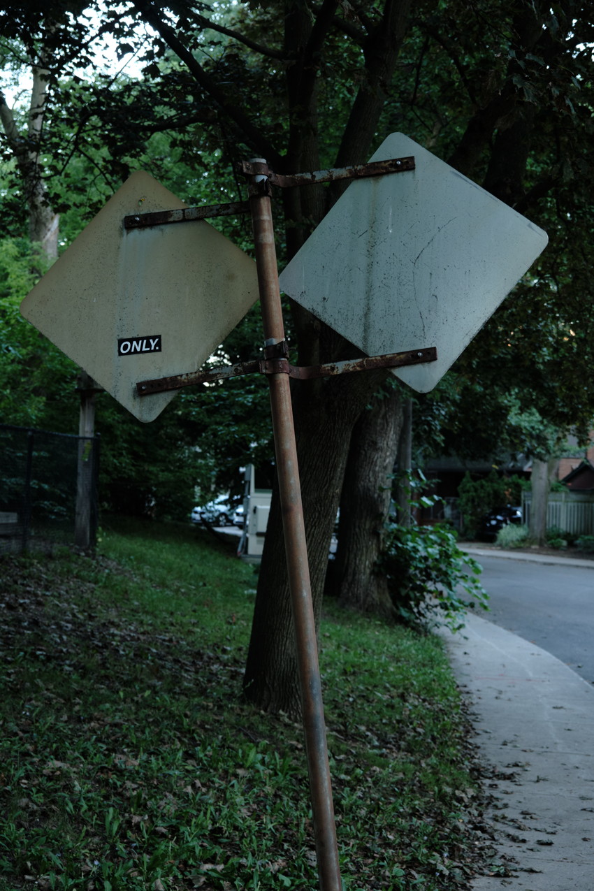 Two weathered diamond-shaped road signs mounted back-to-back on a leaning pole beside a tree-lined sidewalk.