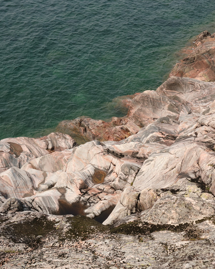 Top view of banded pink-gray coastal rocks descending into green-blue lake, textured rock pools and lichens.
