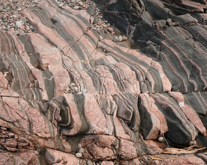 Close-up of layered pink and dark green banded rock with smooth round pebbles scattered in crevices.