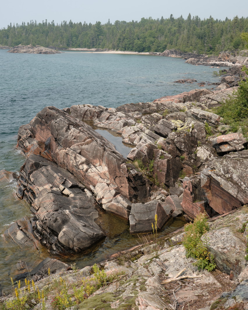 Rocky pink-gray shoreline with tidal pools, calm blue bay, distant sandy cove and dense conifer forest.