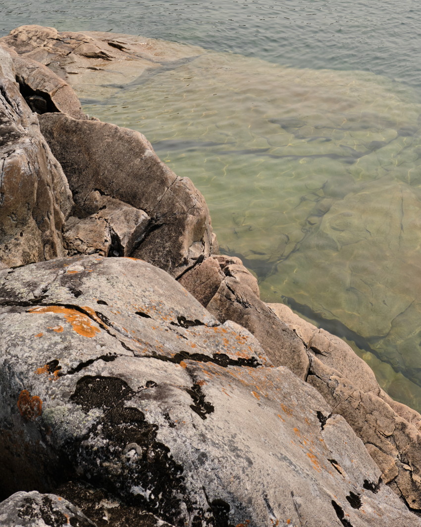 Granite shoreline rocks with orange lichen meeting clear shallow green water revealing submerged rock ledges.