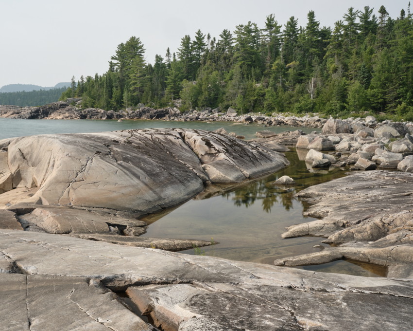 Granite shoreline with smooth weathered rocks, shallow tidal pool reflecting evergreen trees, and distant lake