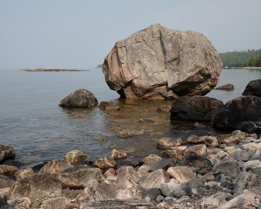 Large boulder at water's edge among smaller rocks, calm water, distant tree-lined shore.