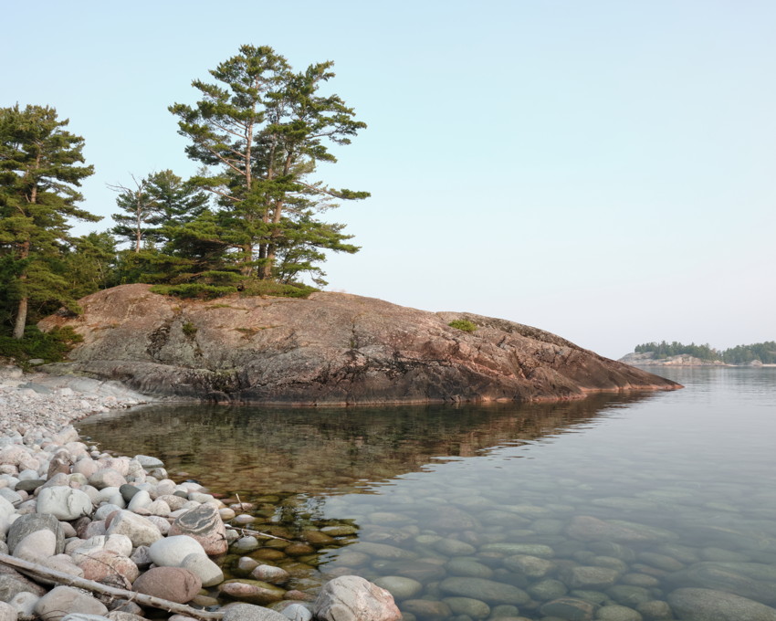 Rocky shoreline with round stones, clear shallow water, granite outcrop topped by pine trees, calm lake and distant tree islands.