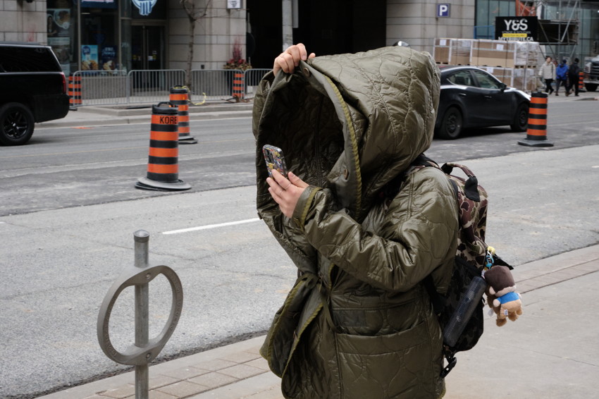 Person in large olive-green quilted coat with hood pulled over face, holding phone, backpack with plush keychain on city sidewalk.