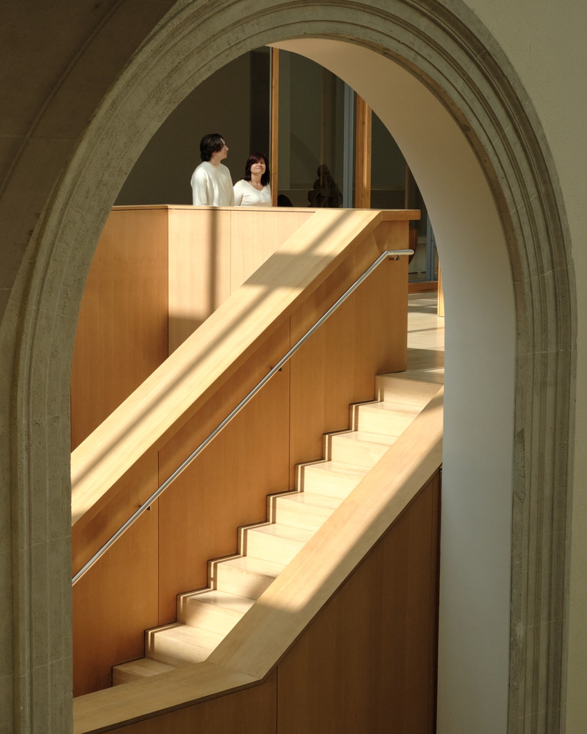 Arch framing wooden staircase with diagonal handrail; two people standing on sunlit upper landing.
