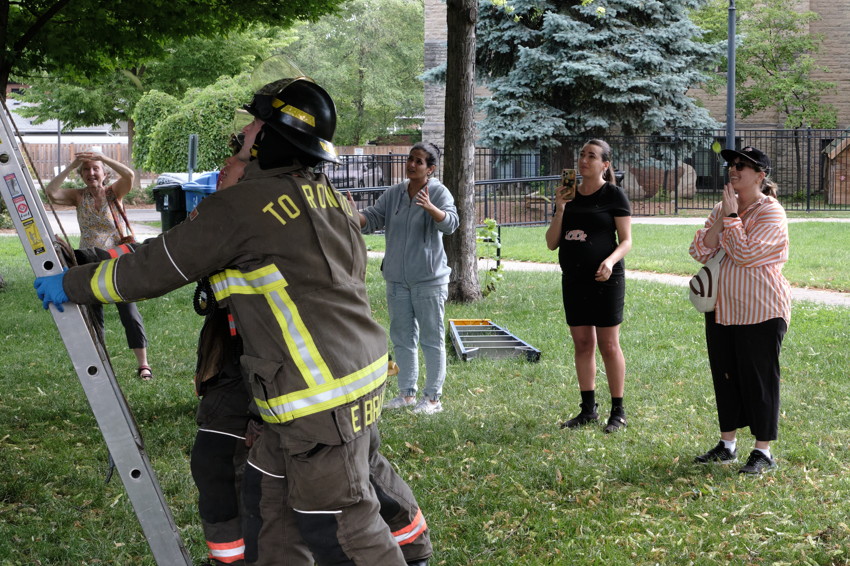 Firefighter in full gear raising a ladder on grass while four women watch nearby under trees in a residential park.