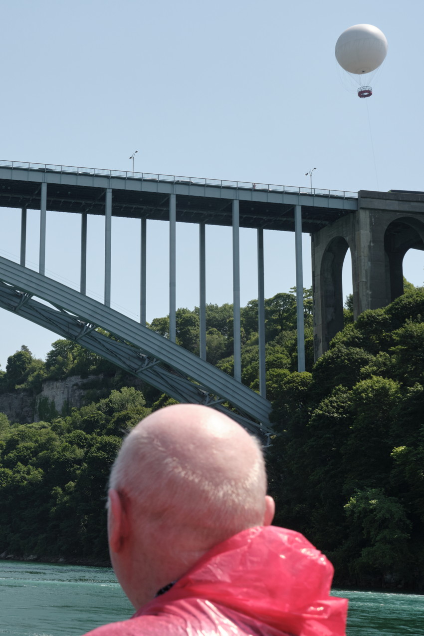 Rear view of bald person in red poncho facing arched steel bridge over river, white tethered balloon overhead.