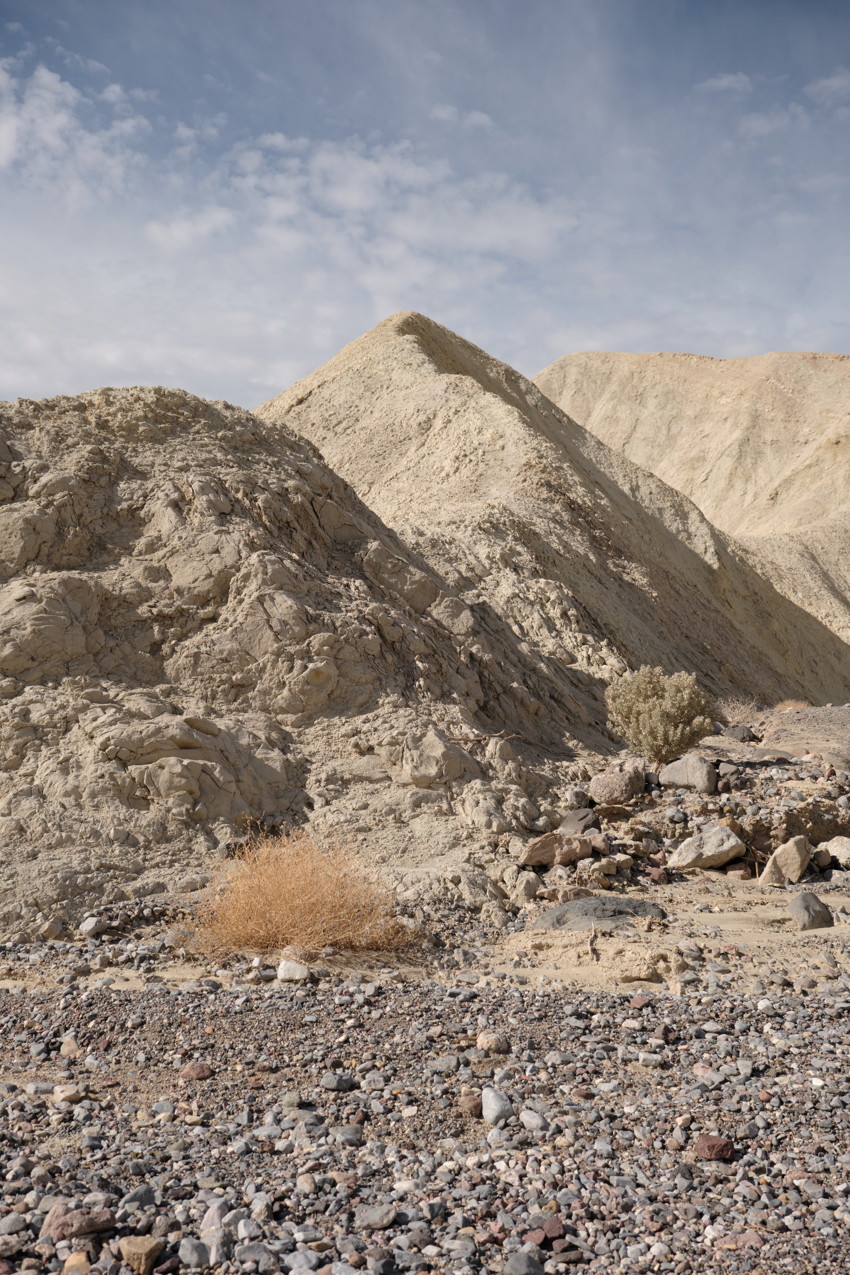 Dry rocky foreground with scattered stones and tumbleweed; eroded pale hills rising under a partly cloudy sky.