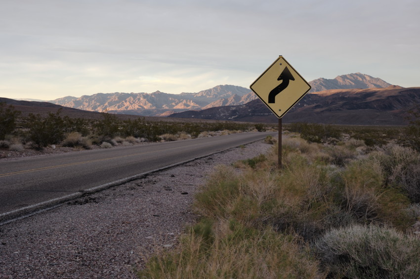 Right-side winding road sign beside two-lane desert road leading toward distant mountains at dusk.