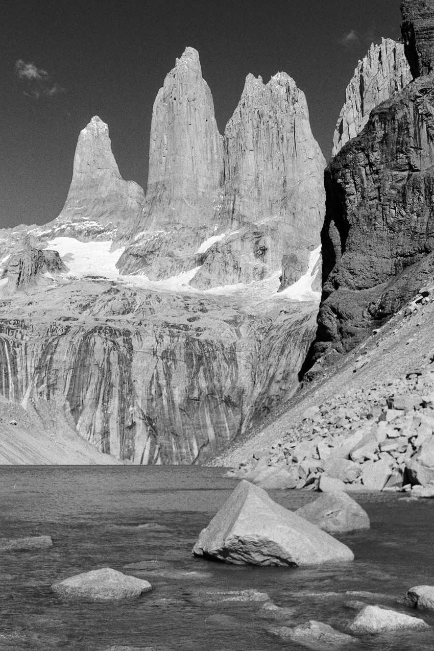 Black-and-white view of three granite towers rising above a glacial basin, lake and boulders in foreground.