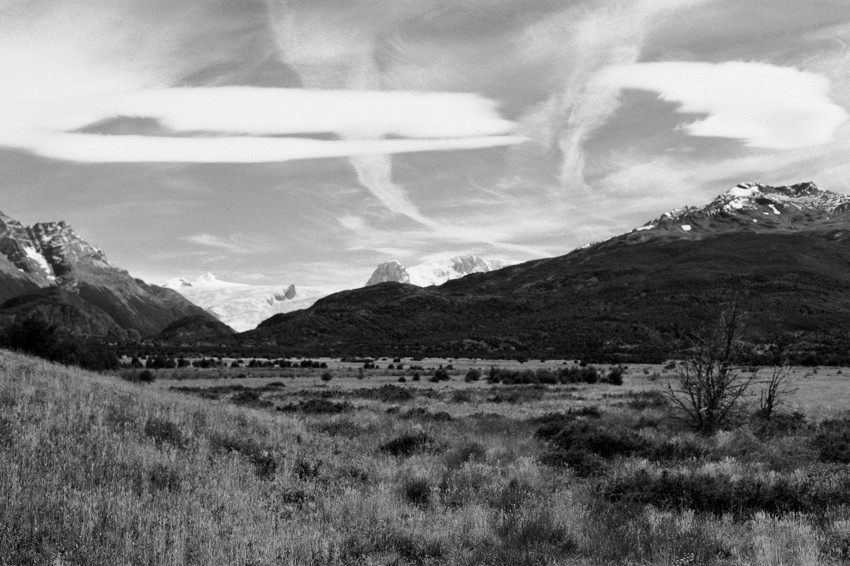 Black-and-white wide landscape: grassy foreground, scattered shrubs, mountain ridge and peaks, large lenticular clouds above.