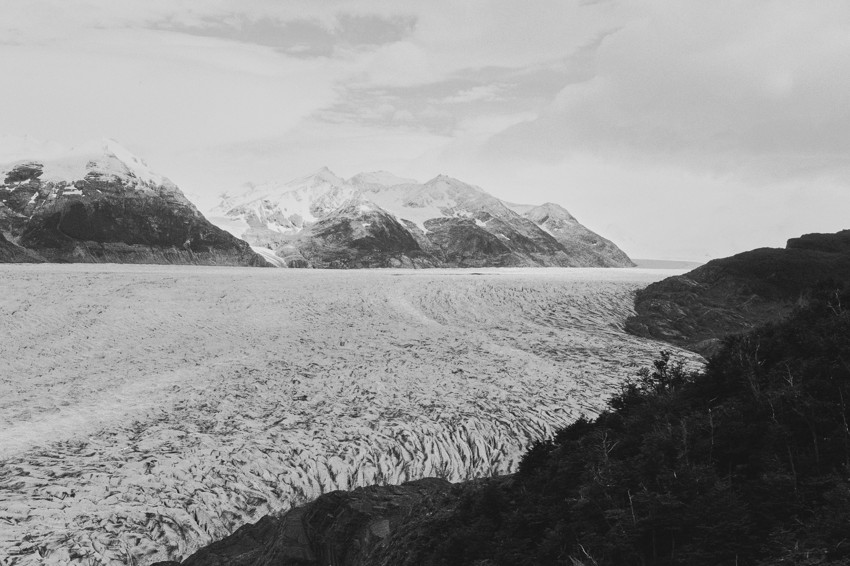 Wide glacier flowing toward distant snow-capped mountains, dark forested ridge in right foreground, cloudy sky.
