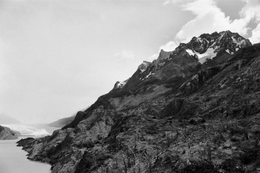 Black-and-white panorama of rugged, snow-capped mountains, glacier at left, rocky sloping foreground.